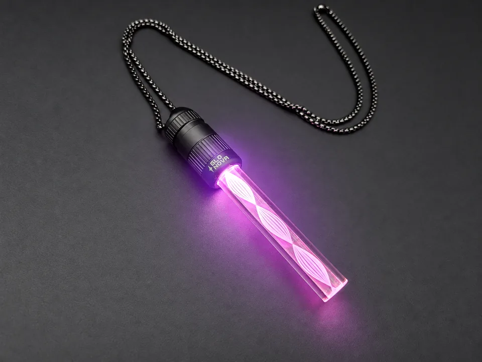 Astral Bloom pendant lying on charcoal with its steel ball chain, a pink spiral of light winding the length of the clear rod