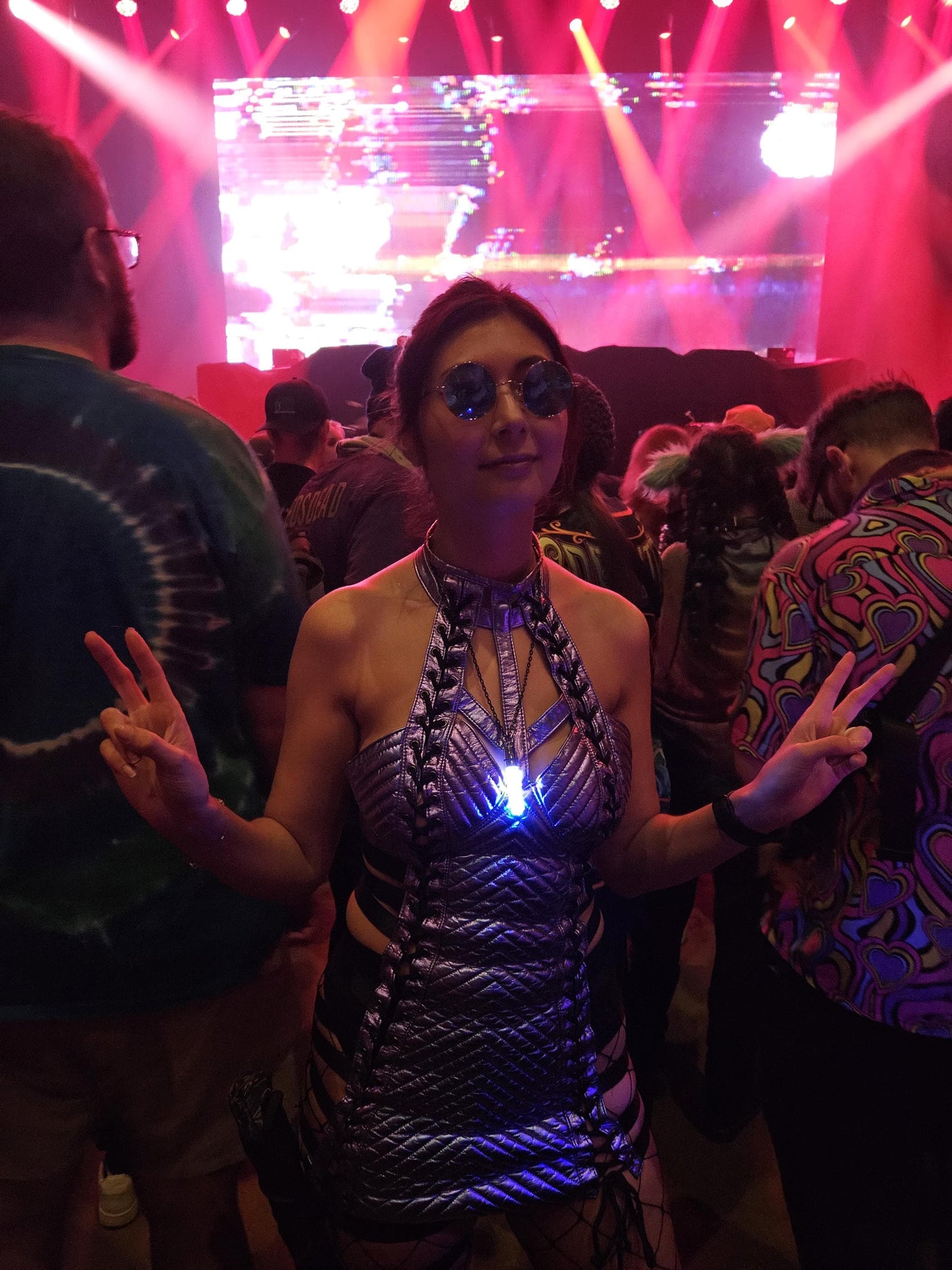 Festival-goer wearing a glowing Glo Nova LED pendant in a silver rave outfit at a concert, surrounded by colorful crowd lights—perfect for raves, EDM events, and cosplay fashion.