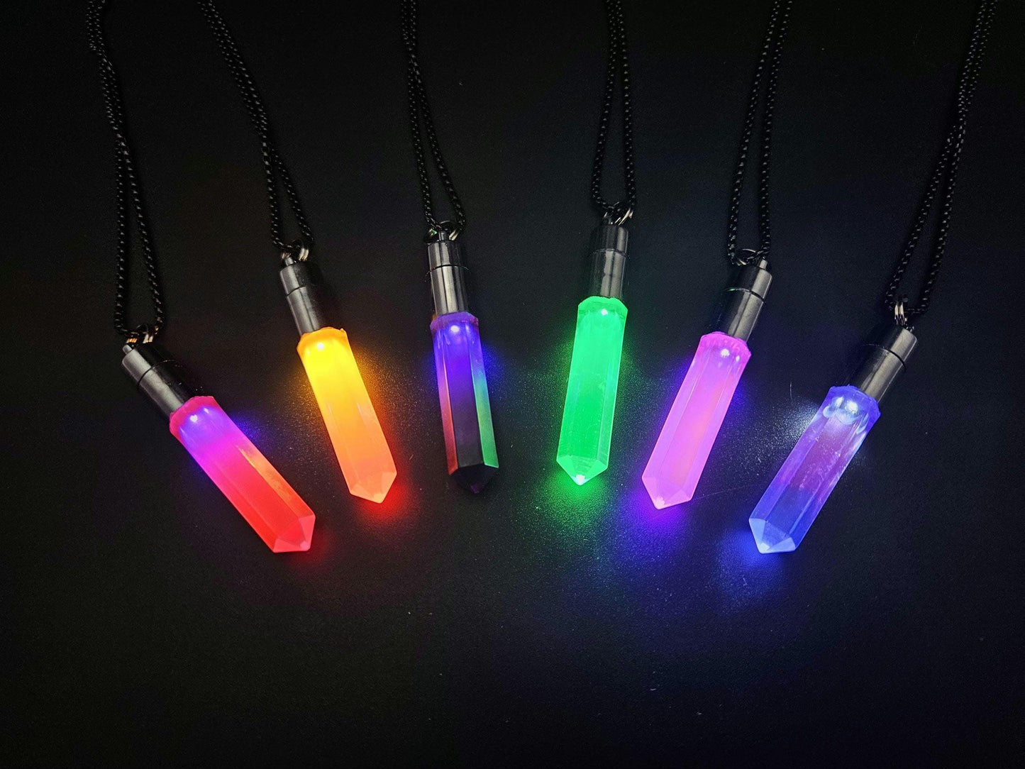 LED glow crystal pendants in assorted colors (red, orange, green, pink, and blue) on black chains, displayed on a dark background — handmade light-up rave jewelry by Glo Nova.